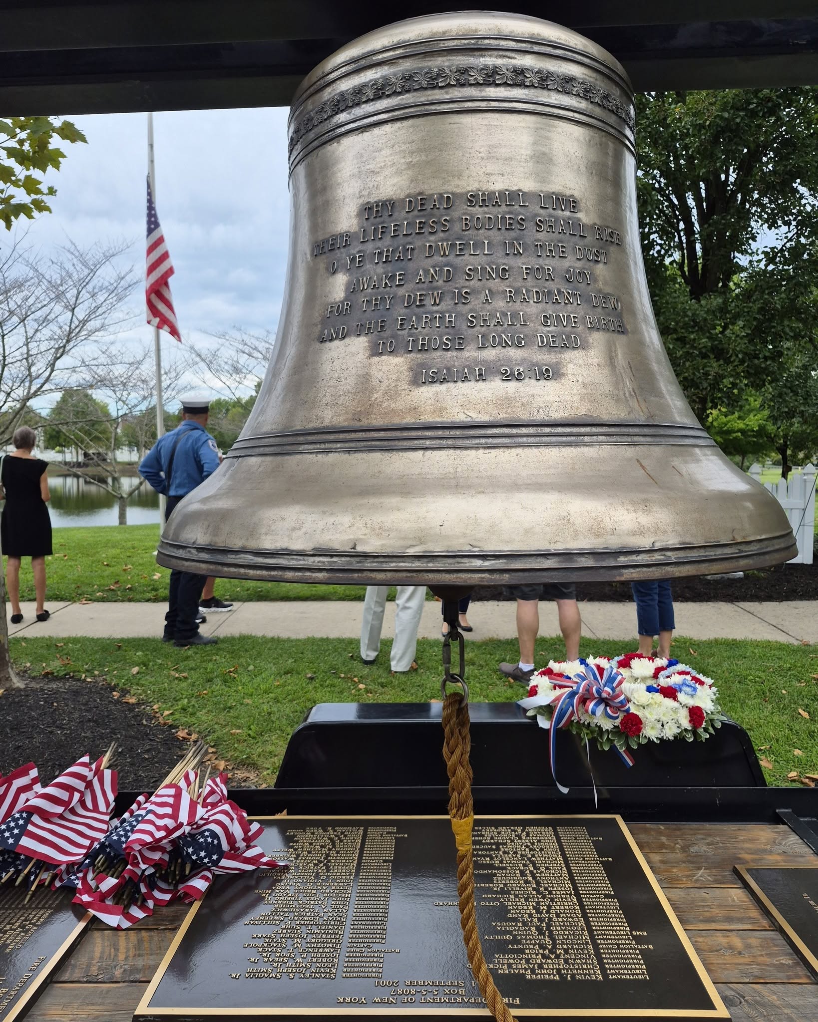 A memorial bell inscribed with Isaiah 26:19, with a red, white and blue wreath and small American flags beside the bronze plaques naming the firefighters lost on September 11, 2001